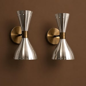 Op de afbeelding: Een paar wandlampen met dubbelconische zilverkleurige metalen kappen met geperforeerde randen. Elke armatuur is gemonteerd op een ronde messingkleurige muurplaat, verbonden door een centraal cilindrisch messingkleurig tussenstuk.