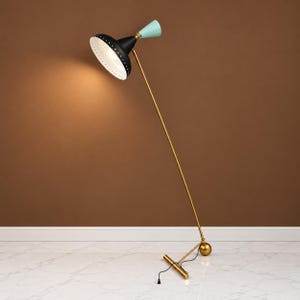 May include: A floor lamp featuring a long, thin gold-toned metal stand and a base with a spherical accent. The lamp head is a black, wide-brimmed shade with a white interior and a small, light blue conical top piece, angled to provide illumination.