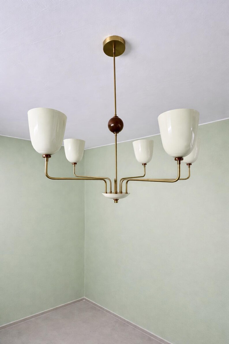 May include: A mid-century modern chandelier with five off-white, tulip-shaped shades. The fixture has brass arms and a dark brown wooden sphere. The ceiling mount is brass. The light fixture is suspended from the ceiling.