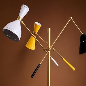 May include: A multi-arm floor lamp featuring a central brass pole with three adjustable conical shades in white, yellow, and black. Each shade has a perforated edge detail. The lamp includes three matching colored handles for positioning the arms.