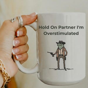May include: White ceramic mug with a handle featuring the text "Hold On Partner I'm Overstimulated." The mug has a cartoon illustration of a frog dressed as a cowboy holding a gun. The mug is held by a person.