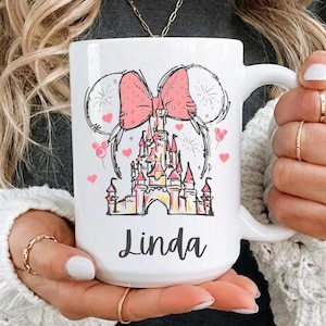 May include: White ceramic mug featuring a pink bow, Mickey Mouse ears, and a castle design. The mug has the name "Linda" printed in black script. Pink hearts and fireworks complete the design. A perfect gift for Disney fans.