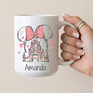 May include: White ceramic mug featuring a pink bow, mouse ears, and a castle design. The mug has the name "Amanda" printed on it. Pink hearts are scattered around the design.
