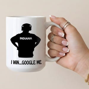 May include: White ceramic mug featuring a black silhouette of a person in a helmet with the word "INDIANA" and the text "I WIN...GOOGLE ME". The mug has a large handle and is held by a hand.