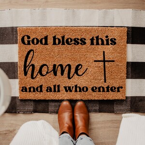May include: A rectangular doormat with the text "God bless this home and all who enter" in black script and a cross symbol. The mat is brown and sits on a black and white checkered rug. Brown boots and blue jeans are visible.