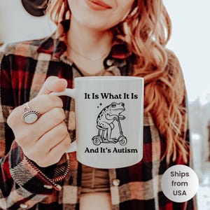 May include: White ceramic mug featuring a black and white illustration of a frog riding a scooter. The mug has the text "It Is What It Is" above the frog and "And It's Autism" below. The mug is held by a person.