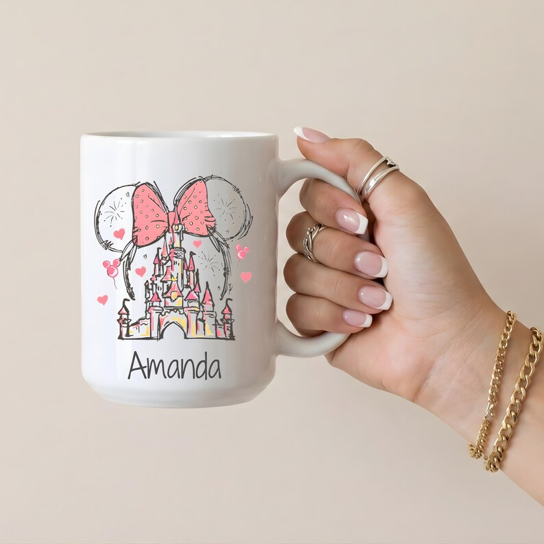 May include: White ceramic mug featuring a pink bow, mouse ears, and a castle design. The mug has the name "Amanda" printed on it. Pink hearts are scattered around the design.