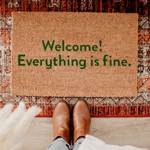 May include: A rectangular doormat made of natural coir fiber. The mat features the text "Welcome! Everything is fine." in green lettering. The mat is placed on a patterned rug, with a person's feet and brown boots visible.