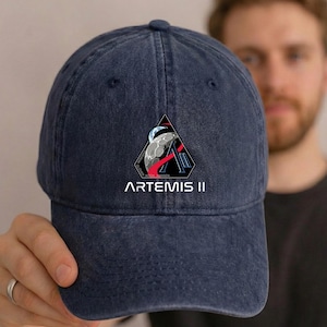 May include: A navy blue baseball cap featuring the Artemis II mission logo. The logo is a black triangle with a stylized "A" and a depiction of the moon and Earth. The text "ARTEMIS II" is printed below the logo in white.