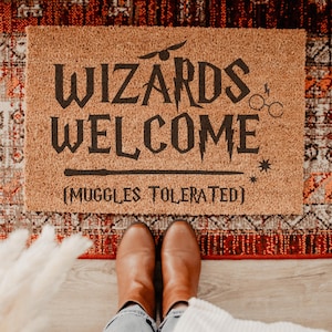 May include: A rectangular coir doormat with the words "WIZARDS WELCOME" in black, along with the phrase "(MUGGLES TOLERATED)". The mat features a lightning bolt, glasses, and a star design. Brown boots and jeans are visible.