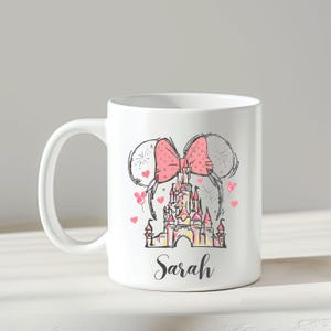 Puede incluir: Taza de cer&aacute;mica blanca con un lazo rosa, orejas de rat&oacute;n, un castillo y corazones. El nombre "Sarah" est&aacute; escrito en una fuente de escritura debajo de la ilustraci&oacute;n del castillo. El dise&ntilde;o es de estilo caprichoso y esbozado.