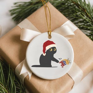 May include: A white ceramic Christmas ornament featuring a black cat wearing a Santa hat. The cat is reaching for a cartoon elephant wearing a crown. The ornament hangs from a gold string and is set against a wrapped gift.