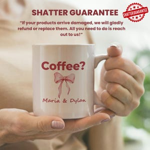Puede incluir: Una taza de cer&aacute;mica blanca con el texto "Coffee?" en borgo&ntilde;a, un lazo de acuarela y los nombres "Maria & Dylan". La taza est&aacute; sostenida por una persona. La imagen tambi&eacute;n incluye un sello de "Shatter Guarantee".