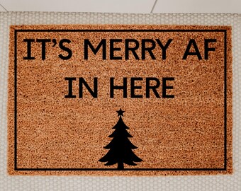May include: A rectangular doormat with a brown coir surface and a black border. The text "IT'S MERRY AF IN HERE" is printed in black, along with a black Christmas tree graphic. The mat is designed for holiday decor.
