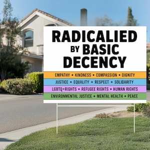 May include: A white sign with the text "RADICALIED BY BASIC DECENCY" in bold black letters. Below, colorful horizontal bars list values like "EMPATHY," "JUSTICE," and "LGBTQ+ RIGHTS." The sign is displayed on a grassy lawn.