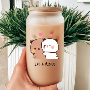 May include: A clear glass tumbler with a bamboo lid, filled with a creamy beverage and ice cubes. The tumbler features a cartoon illustration of two hugging bears with pink hearts above them. The names "Leo & Aisha" are printed below.
