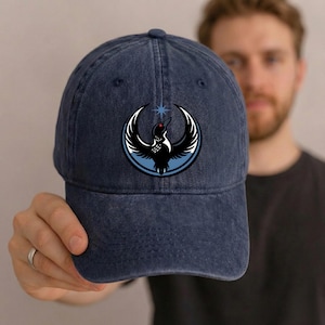 May include: A dark blue baseball cap with a curved brim. The cap features a circular emblem with a black bird design, white wings, and a red eye, set against a light blue circle. A star is above the bird.