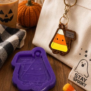May include: A purple silicone mould featuring a candy corn character with a sign that reads I HATE YOU TOO, alongside a matching wooden keychain of the same character, and a canvas bag with a ghost graphic and the text STAY SPOOKY.