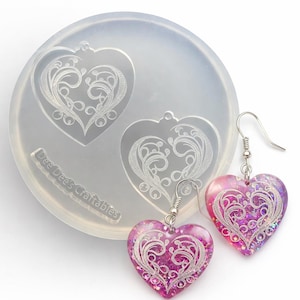 May include: A clear silicone mold with two heart-shaped cavities, each featuring an ornate, etched design. Two finished heart-shaped earrings with silver hooks are displayed, showcasing a pink and purple glittery resin with the same etched design. The mold has the text "Dee Dee's Craftables".