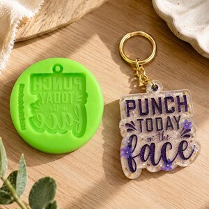 Può includere: Uno stampo in silicone verde con il testo "PUNCH TODAY in the face" al contrario. Accanto, un portachiavi trasparente con lo stesso testo in glitter viola e oro, con piccoli accenti floreali viola.