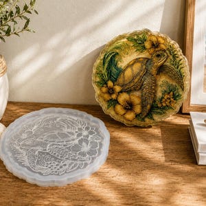 Pode incluir: Um molde de silicone redondo e translúcido com uma tartaruga marinha em relevo rodeada por flores de hibisco e folhagem, acompanhado por um porta-copos de resina circular acabado com um design detalhado de tartaruga marinha e flores amarelas.