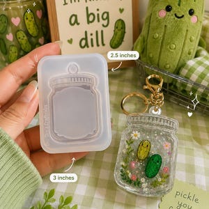 May include: A rectangular silicone mold for a jar-shaped keychain, measuring 2.5 inches by 3 inches, alongside a finished clear resin keychain containing two small green pickle figures, tiny white and pink flowers, and silver glitter.