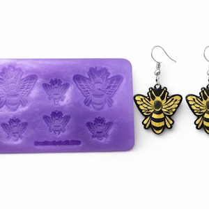 May include: A purple silicone mold with six bee impressions and a pair of bee-shaped earrings. The earrings have black outlines, gold wings, and silver hooks. The mold is rectangular with rounded corners.