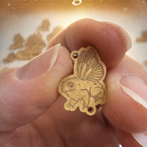 Könnte beinhalten: Ein kleiner, goldfarbener Anhänger in Form eines Frosches mit Schmetterlingsflügeln wird hochgehalten. Der Anhänger hat zwei Löcher oben und ist mit Details graviert. Im Hintergrund steht "Coming Soon".