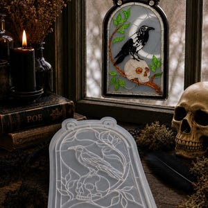 May include: A translucent silicone mold featuring an arched design with a raven perched on a human skull amidst leafy branches, paired with a finished stained glass-style hanging panel depicting the same raven and skull scene in black, white, and green.
