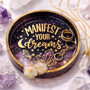 May include: A round tray with a gold rim and a galaxy-themed interior, featuring the text "MANIFEST YOUR DREAMS". The tray holds rings and earrings, with gold necklaces and crystals nearby. Amethyst and clear quartz crystals are scattered around.