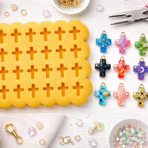 May include: A collection of colorful cross-shaped resin charms with gold-colored jump rings, alongside a yellow cross-shaped mold. Various colors and glitter are visible in the charms. Additional jewelry-making supplies are also present.