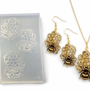 Puede incluir: Un conjunto de joyas con temática de abeja. El conjunto incluye un collar y pendientes con detalles de abeja en metal dorado y negro y amarillo. También hay un molde con diseños de abeja y panal.