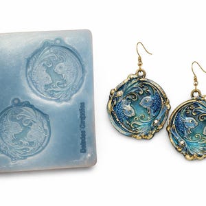 May include: A pair of ornate, blue and gold earrings featuring a fish design, next to a blue mold with the same design. The earrings have a detailed, vintage aesthetic with a gold-colored hook.
