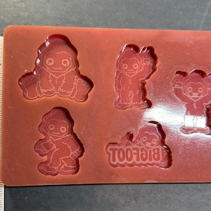 Puede incluir: Un molde de silicona rojo con cinco diseños de Bigfoot. Las figuras de Bigfoot están en varias poses, incluyendo sentado, saludando y de pie. La palabra "BIGFOOT" también está moldeada al revés. Una regla está al lado del molde.