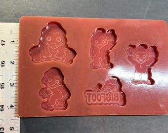 Könnte beinhalten: Eine rote Silikonform mit fünf Bigfoot-Designs. Die Bigfoot-Figuren befinden sich in verschiedenen Posen, darunter sitzend, winkend und stehend. Das Wort "BIGFOOT" ist ebenfalls umgekehrt geformt. Ein Lineal befindet sich neben der Form.