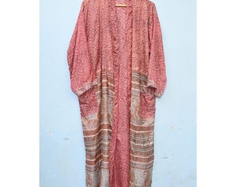 Op de afbeelding: Lange, openvallende kimono in roze en bruine tinten, hangend aan een houten hanger. De kimono heeft een paisley patroon langs de onderkant en mouwen, met een bloemenmotief dat de rest van het kledingstuk bedekt. De kimono is gemaakt van een zijdeachtig materiaal.