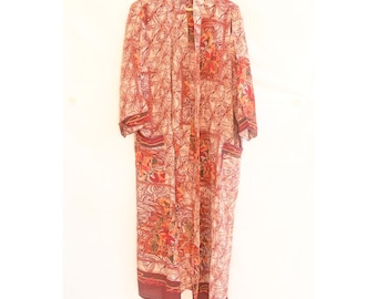 Op de afbeelding: Een lange, openvallende kimono met een bloemen- en geometrisch patroon in tinten rood, oranje en crème. De kimono heeft lange mouwen, zakken en een bordeauxrode rand. Hij hangt aan een houten kleerhanger.