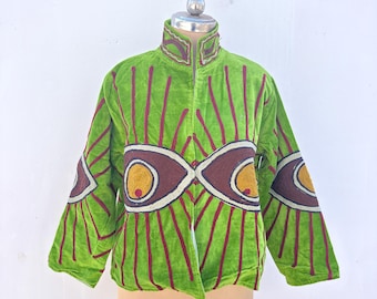 May include: A green velvet jacket with a high collar and long sleeves. The jacket features a pattern of vertical red stripes and large eye-shaped designs in brown, yellow, and white. The jacket is displayed on a mannequin.