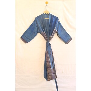 Puede incluir: Una bata azul con mangas largas y cierre de lazo. La bata tiene un borde estampado en tonos marrones y blancos. La bata está colgada de una percha de madera sobre un fondo blanco.