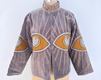 Puede incluir: Chaqueta de terciopelo gris con cuello alto y mangas largas. La chaqueta presenta patrones bordados de rayos de sol y grandes diseños de ojos en tonos dorados, naranjas y blancos. La chaqueta se exhibe en un maniquí.