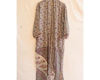Op de afbeelding: Een lange, openstaande kimono met een bloemenpatroon in tinten paars, groen en crème. De kimono heeft lange mouwen, zakken en een contrasterende rand aan de zoom. Hij hangt aan een houten hanger tegen een neutrale achtergrond.
