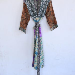 May include: A patterned robe with long sleeves hangs against a white wall. The robe features a mix of floral and paisley designs in shades of green, orange, purple, and blue. A matching belt is tied in the middle.