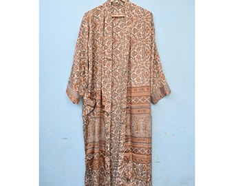 Op de afbeelding: Een lange, openstaande kimono met een bloemenpatroon in bruin, beige en grijstinten. De kimono heeft lange mouwen en twee zakken. Hij hangt aan een houten hanger tegen een lichtblauwe muur.