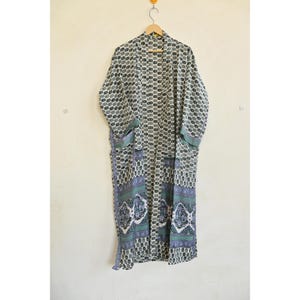 May include: A long, open-front kimono robe with a repeating geometric pattern in shades of gray and white. The robe has a contrasting border with a floral design in blue and green. It is hanging on a wooden hanger.