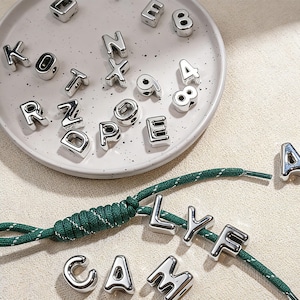 May include: Shiny silver metal alphabet and number beads, some on a white plate, others strung on a green and white cord. The letters spell "CAM LYF A". The beads are reflective.