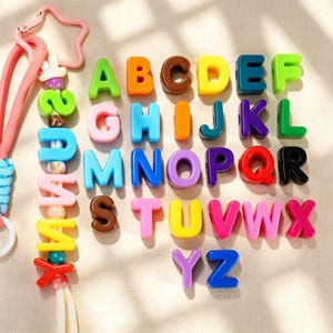Può includere: Una collezione di perline colorate in plastica con le lettere dell'alfabeto dalla A alla Z in varie tonalità vivaci. Un lungo cordino è decorato con una serie di queste perline a lettera, perline rotonde più piccole e una chiusura a stella rosa.