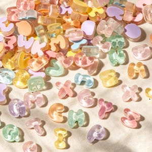 Puede incluir: Una colección de pequeñas cuentas con letras del alfabeto, translúcidas y opacas, en varios colores pastel que incluyen rosa, amarillo, verde, naranja y morado. Las cuentas tienen formas redondeadas y gruesas, dispersas de forma aleatoria.