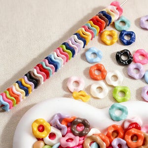 Könnte beinhalten: Eine Sammlung bunter, donut-förmiger Perlen in verschiedenen Farben wie Rosa, Blau, Orange und Grün. Einige Perlen sind auf einem Draht aufgefädelt, andere liegen um eine weiße Schale herum. Die Perlen haben eine glänzende Oberfläche.