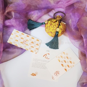 May include: A gold pouch with a floral design, teal tassels, and gold details. Several white cards with orange cloud patterns and text are arranged around the pouch. The background features a sheer purple fabric.