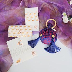 May include: A blue and red floral patterned pouch with a gold handle and blue tassels, alongside white cards featuring cloud and floral designs. The cards display text and a logo. The background is a purple fabric.
